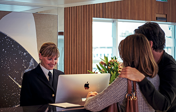 A couple at a reception desk being assisted by a smiling woman looking at a computer screen.