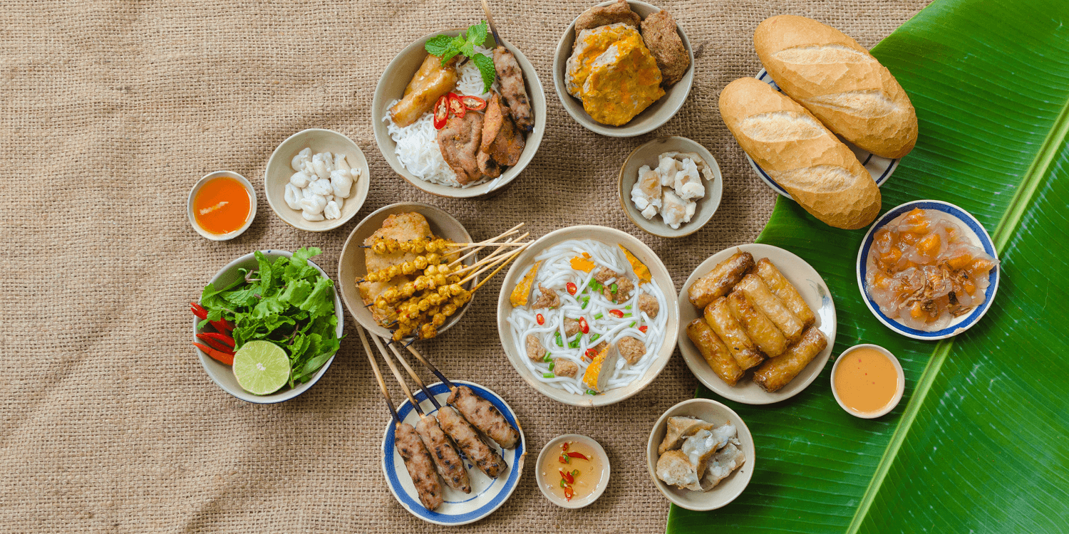 Top-down view of assorted traditional Vietnamese dishes, including noodle soups, grilled meat skewers, spring rolls, fresh herbs, and baguettes on a woven mat.