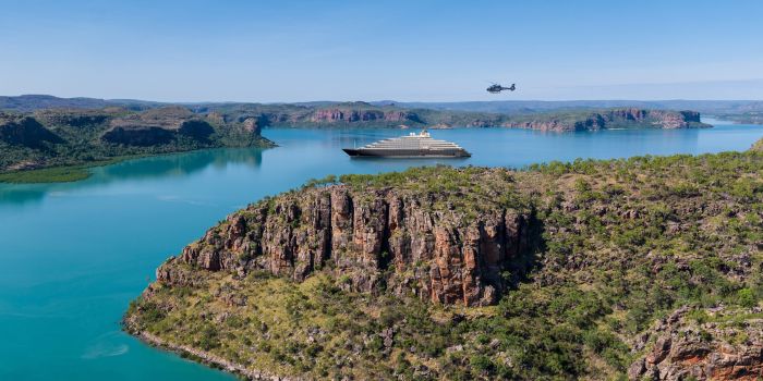 Scenic Eclipse cruising through turquoise waters in the Kimberley, Australia, with a helicopter flying above rugged cliffs